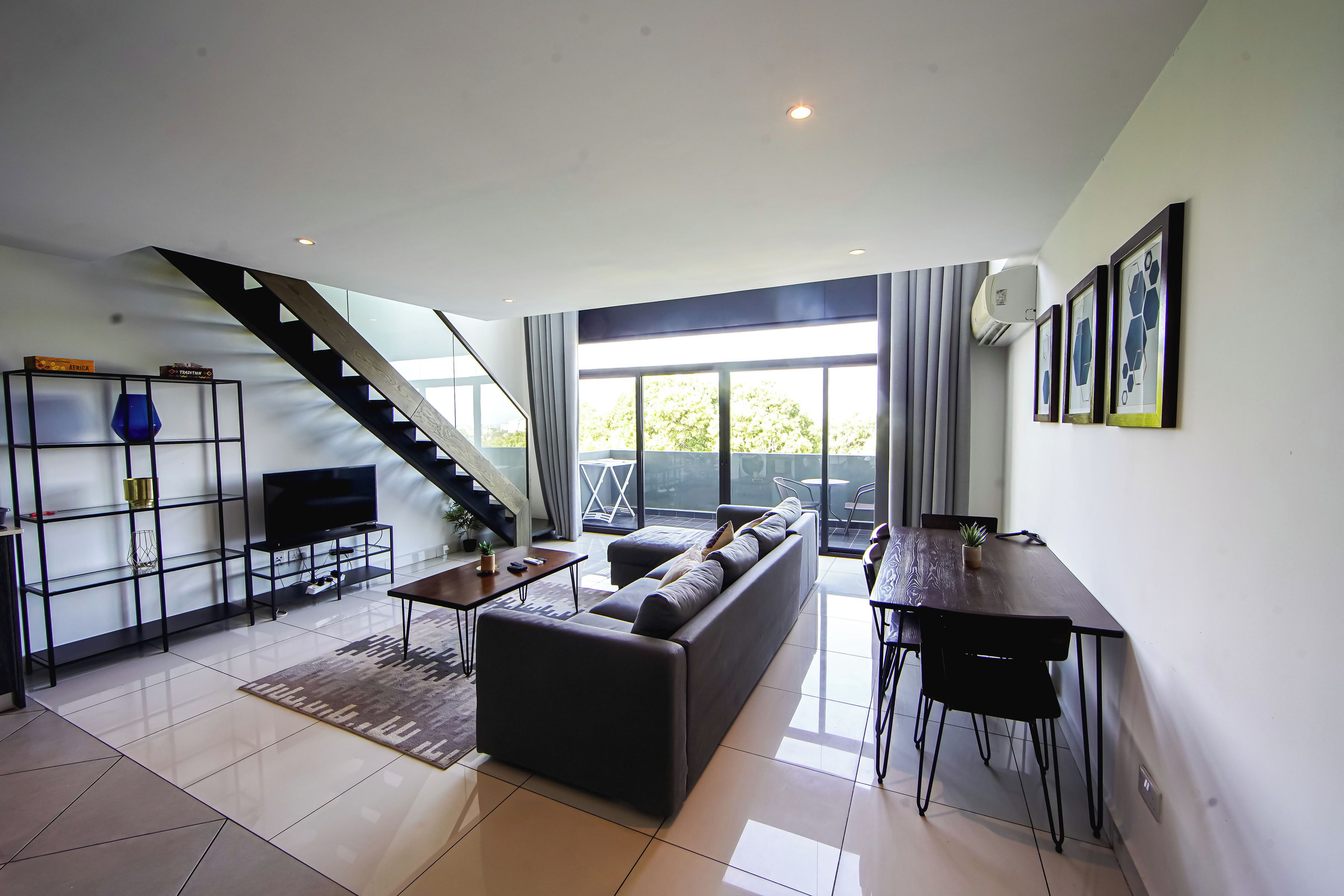Una sala de estar moderna con un sofá, una mesa de centro y un televisor sobre un mueble. Se ve una escalera que conduce a un balcón con vistas.