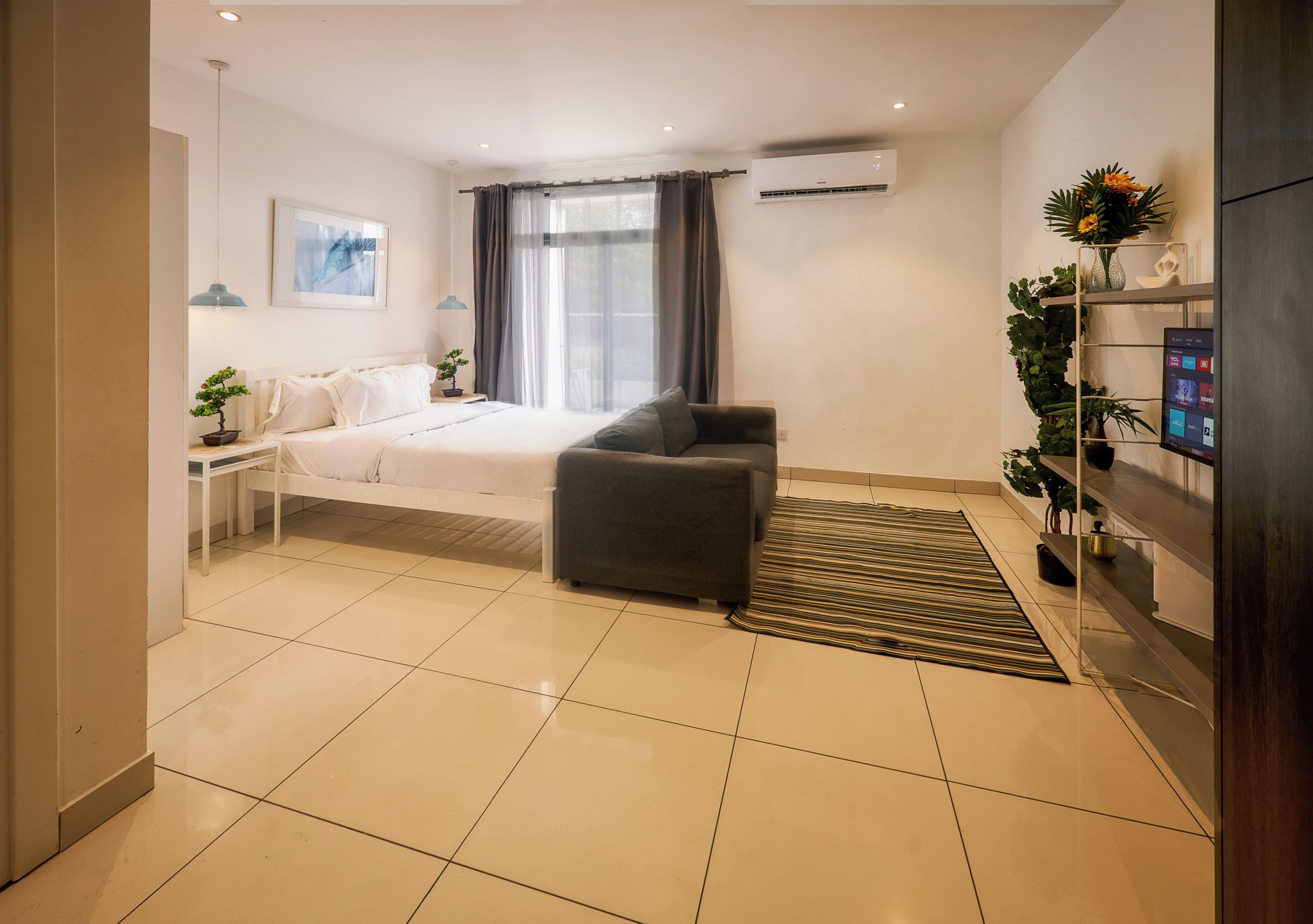 A modern hotel room with a bed, a sofa, a TV, a shelf with plants, and an air conditioning unit.