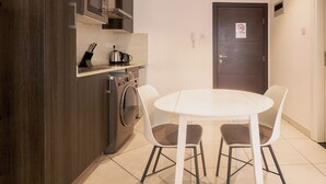 A compact kitchen with a microwave, kettle, and washing machine, a white round table with chairs, and a door with a no-smoking sign. - Peravi at Embassy Gardens  (Accra)