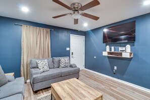 Ein Wohnzimmer mit grauem Sofa, einem Holztisch, einem Deckenventilator und einem an der Wand befestigten Fernseher.