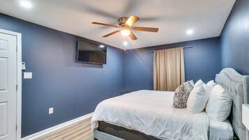 Une chambre à coucher avec un lit, un ventilateur de plafond, un téléviseur fixé au mur et une fenêtre avec des rideaux.