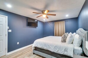 Ein Schlafzimmer mit Bett, Deckenventilator, fest installiertem Fernseher und einem Fenster mit Vorhängen.