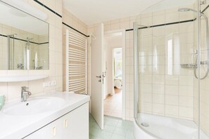 Een badkamer met een douchecabine van glas, een witte wastafel en een deur naar een andere kamer.