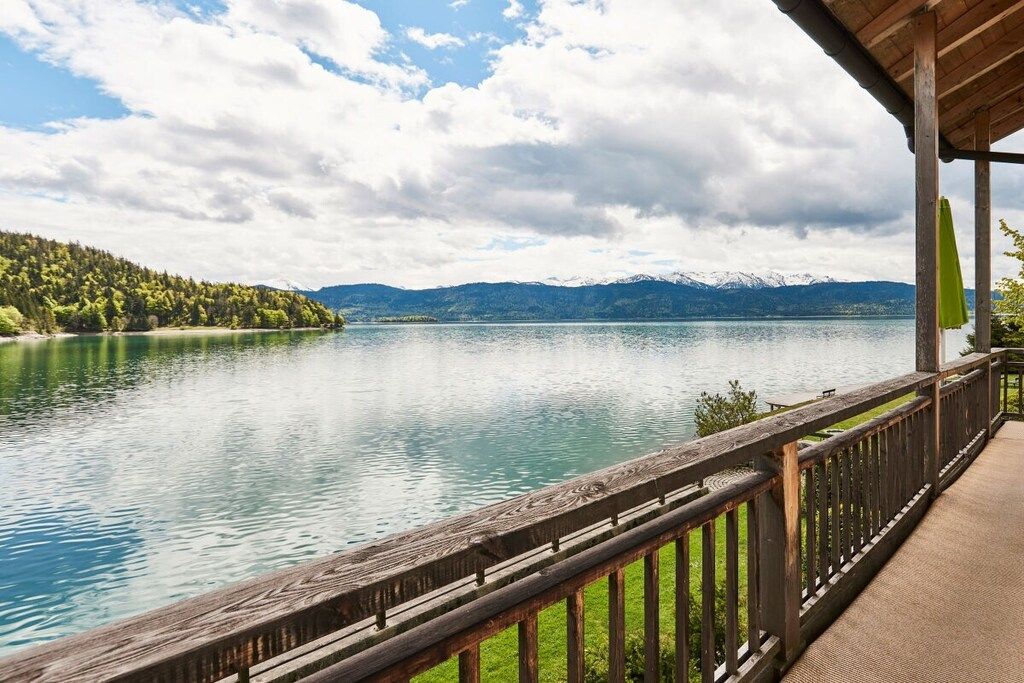 Una terraza de madera da vista a un lago tranquilo con una orilla boscosa y montañas a lo lejos.