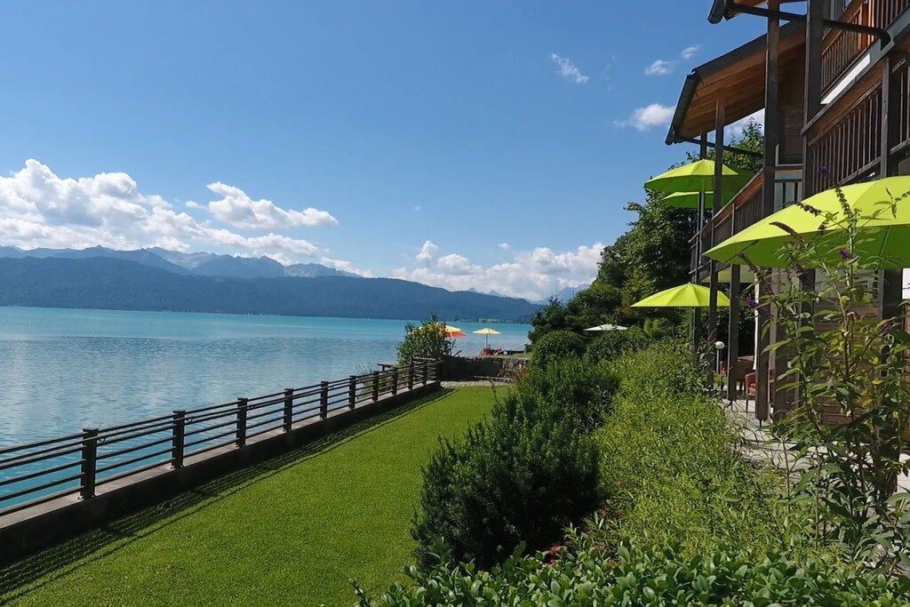 A lakeside hotel with a view of mountains, a grassy lawn, and outdoor umbrellas.