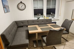 Ein moderner Essbereich mit Holztisch und gepolsterten Stühlen, einer Uhr an der Wand und einem Fenster mit Jalousien.