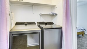 Lavanderia con due lavatrici e un'asciugatrice, uno scaffale sopra gli elettrodomestici e un letto nella stanza adiacente.
