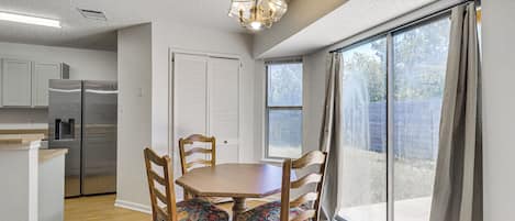 A dining area with a wooden table and chairs, a kitchen with a refrigerator, and a large window with curtains.