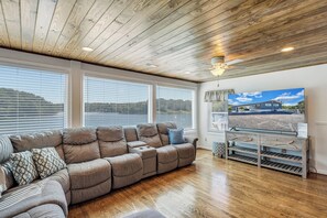 A room with a wooden ceiling, a sectional sofa, a TV on a wooden stand, and large windows with blinds.
