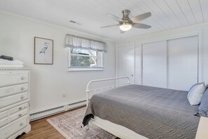 Ein Schlafzimmer mit Bett, Schminktisch, Fenster mit Jalousien, Deckenventilator und einem gerahmten Bild an der Wand.