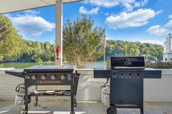 A barbecue grill and a charcoal grill under a covered area with a view of a lake and trees.