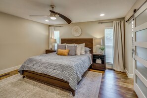 A bedroom with a large bed, wooden headboard, bedside tables, a ceiling fan, and a window with curtains. - Boat Dock & Game Room! Lake Hamilton Home (Hot Springs)