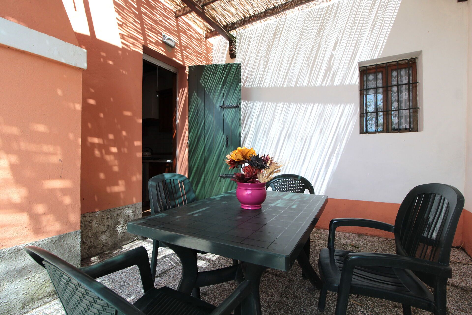 A patio with a table and chairs, a pink vase with flowers, and a green door.