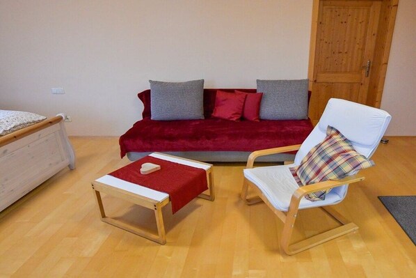 A room with a wooden floor, a red sofa with grey cushions, a white armchair with a plaid cushion, and a small wooden table with a red cloth. - Holiday apartment with 1 bedroom (Salzweg)