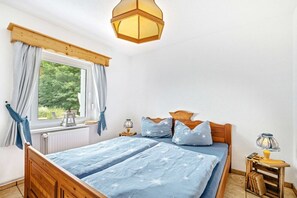 A bedroom with a wooden bed, a window with curtains, a nightstand with a lamp, and a view of greenery outside. - ||---------------------"Haus Waldesruh"------------------|| (Elbingerode)