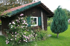 Eine rustikale Holzhütte mit einem grünen Dach, umgeben von einem Garten mit rosa Blumen und einem Nadelbaum.