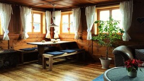 Interior acolhedor de uma cabana com uma mesa de jantar de madeira, bancos, uma planta em vaso e um sofá de couro.