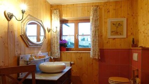 Casa de banho com painéis de madeira, um espelho redondo, uma pia, uma sanita e uma janela com cortinas.