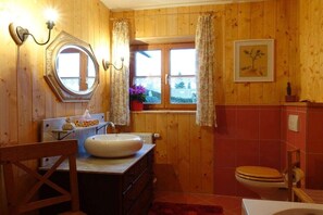 A wooden-paneled bathroom with a round mirror, a sink, a toilet, and a window with curtains. - Morning Dew - Rosenhof-Allgäu (Görisried)