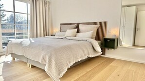 Ein modernes Schlafzimmer mit einem großen Bett, einem Holz-Kopfteil und Blick ins Freie durch ein großes Fenster.
