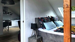 Ein modernes Schlafzimmer mit schrägen Wänden, einem Bett mit floralem Bettzeug, einer Nachttischlampe und Blick auf eine Küche.