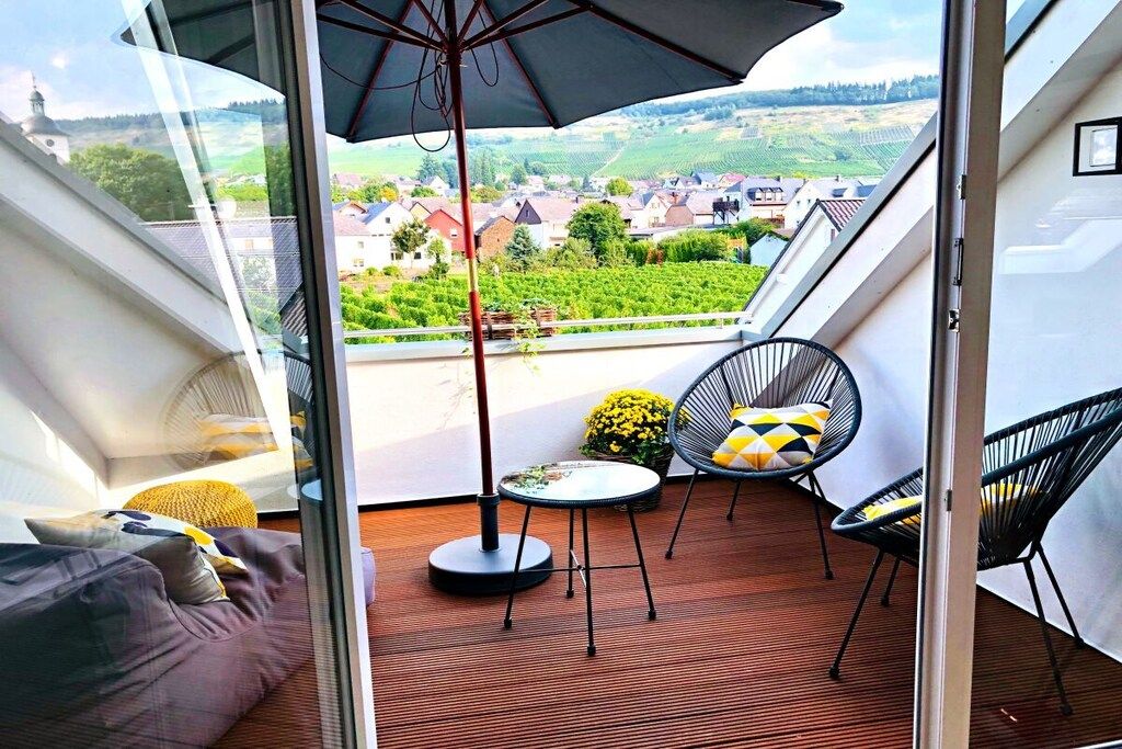 A balcony with a view of a vineyard and houses, a round table, and two chairs.