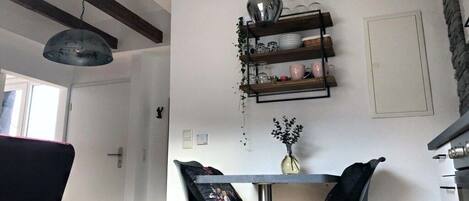 A dining area with a table, two chairs, a hanging light fixture, and a wall-mounted shelf with dishes.
