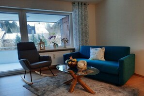 Un salon avec un canapé bleu, un fauteuil à bascule, une table basse en verre et une vue sur un immeuble à l’extérieur.