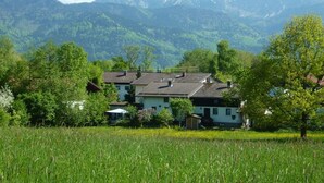 Uma casa com telhado inclinado, rodeada por um campo e árvores, com montanhas ao fundo.