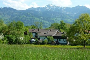 A house with a sloped roof, surrounded by a field and trees, with mountains in the background. - Monika House in Übersee am Chiemsee (Übersee)