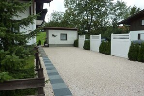 A gravel driveway leading to a white building with a small attached structure, surrounded by greenery and a white fence. - Monika House in Übersee am Chiemsee (Übersee)