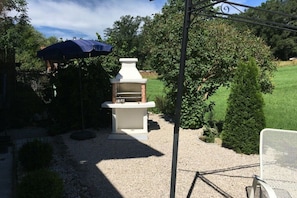 An outdoor barbecue grill with a chimney, surrounded by gravel, trees, and a metal chair. - Monika House in Übersee am Chiemsee (Übersee)