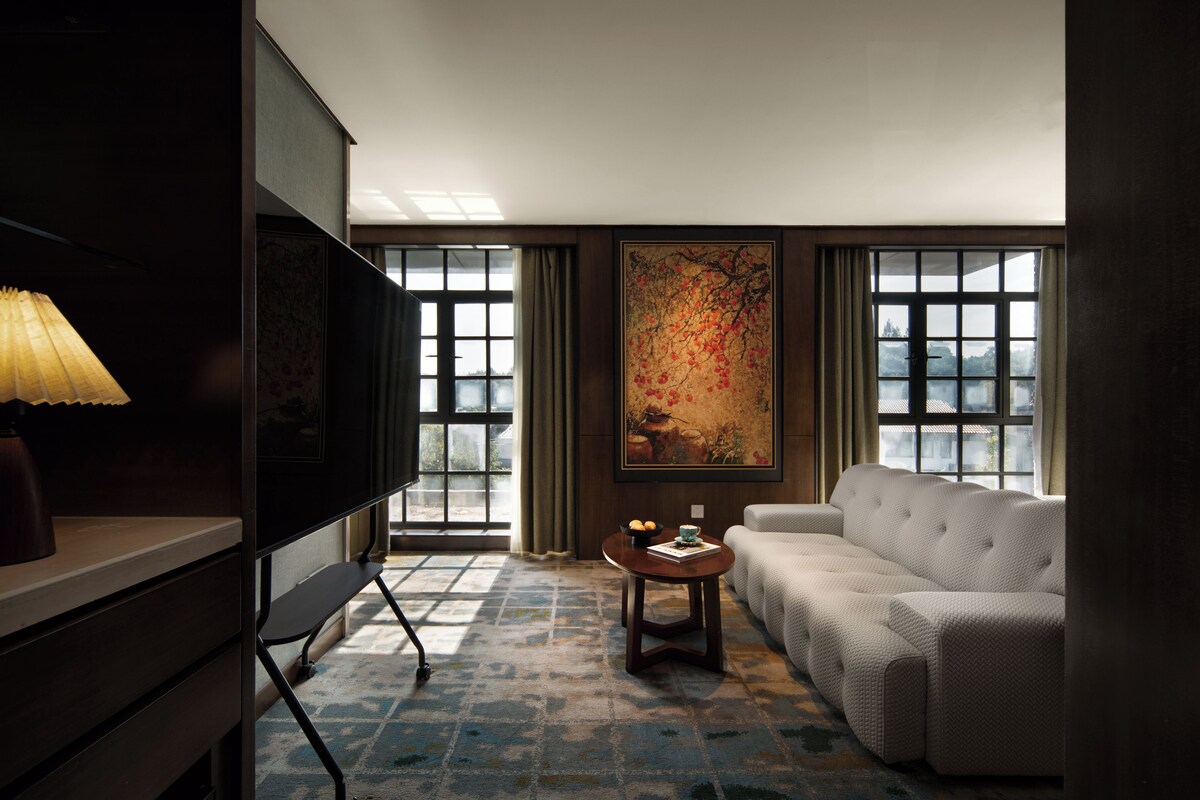 A hotel room with a sofa, a television, a painting, and a view through the windows.