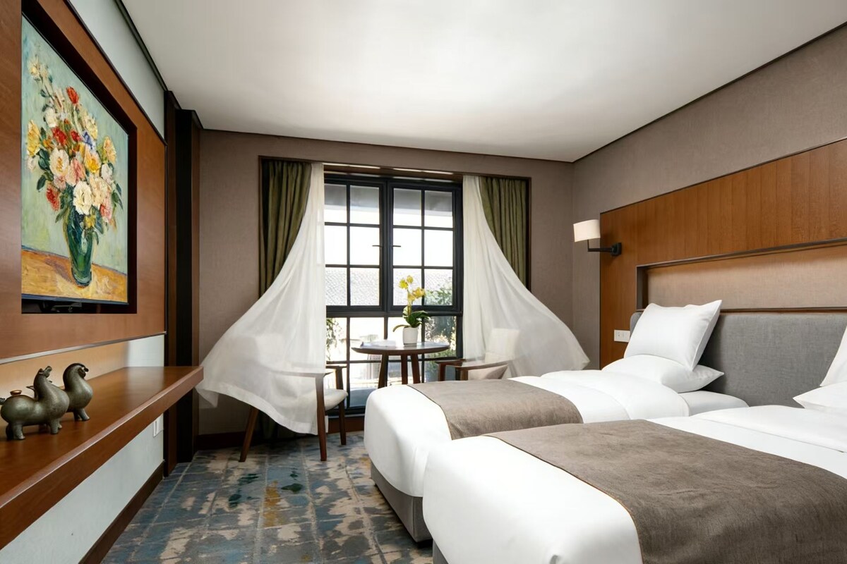 A hotel room with two beds, a desk, a chair, a window with curtains, and a painting on the wall.