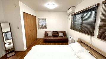 A bedroom with a bed, a wooden wardrobe, a brown sofa, and a window with blinds.