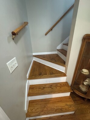 Una escalera con escalones de madera y barandilla, un interruptor de luz en la pared, y un mueble de madera con objetos decorativos.