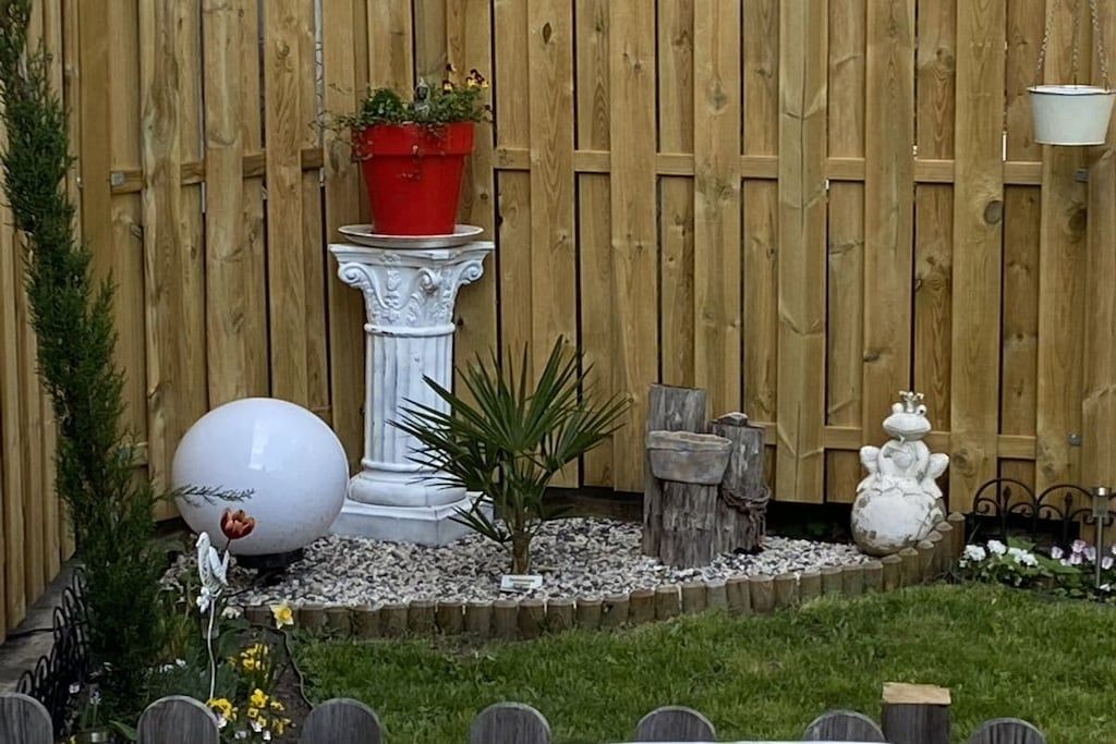 A garden with a wooden fence, a white decorative column, a spherical sculpture, and a red planter with flowers.