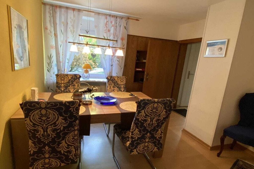 A dining area with a table set for four, patterned chairs, a window with curtains, and a door leading to another room.