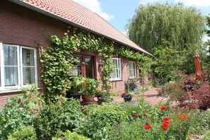 A brick house with a red roof, a garden with various plants and flowers, and a pathway leading to the entrance. - Holiday apartment with 2 bedrooms (Wölpinghausen)