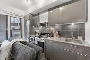Eine moderne Küche mit Herd, Backofen und Spüle, ein graues Sofa mit Decke und ein Fenster mit Blick auf Gebäude.