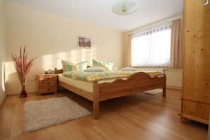 Ein Schlafzimmer mit einem hölzernen Bett, einem Nachttisch, einer Vase mit Trockenpflanzen und einem Fenster mit Vorhängen.