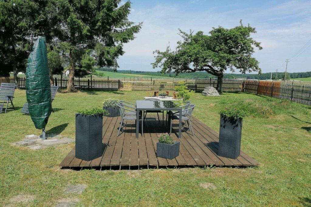 A wooden deck with a table and chairs, surrounded by a fenced area with trees and a grassy field in the background.