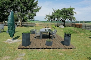 A wooden deck with a table and chairs, surrounded by a fenced area with trees and a grassy field in the background. - Melitta holiday apartment (Heiligenberg)