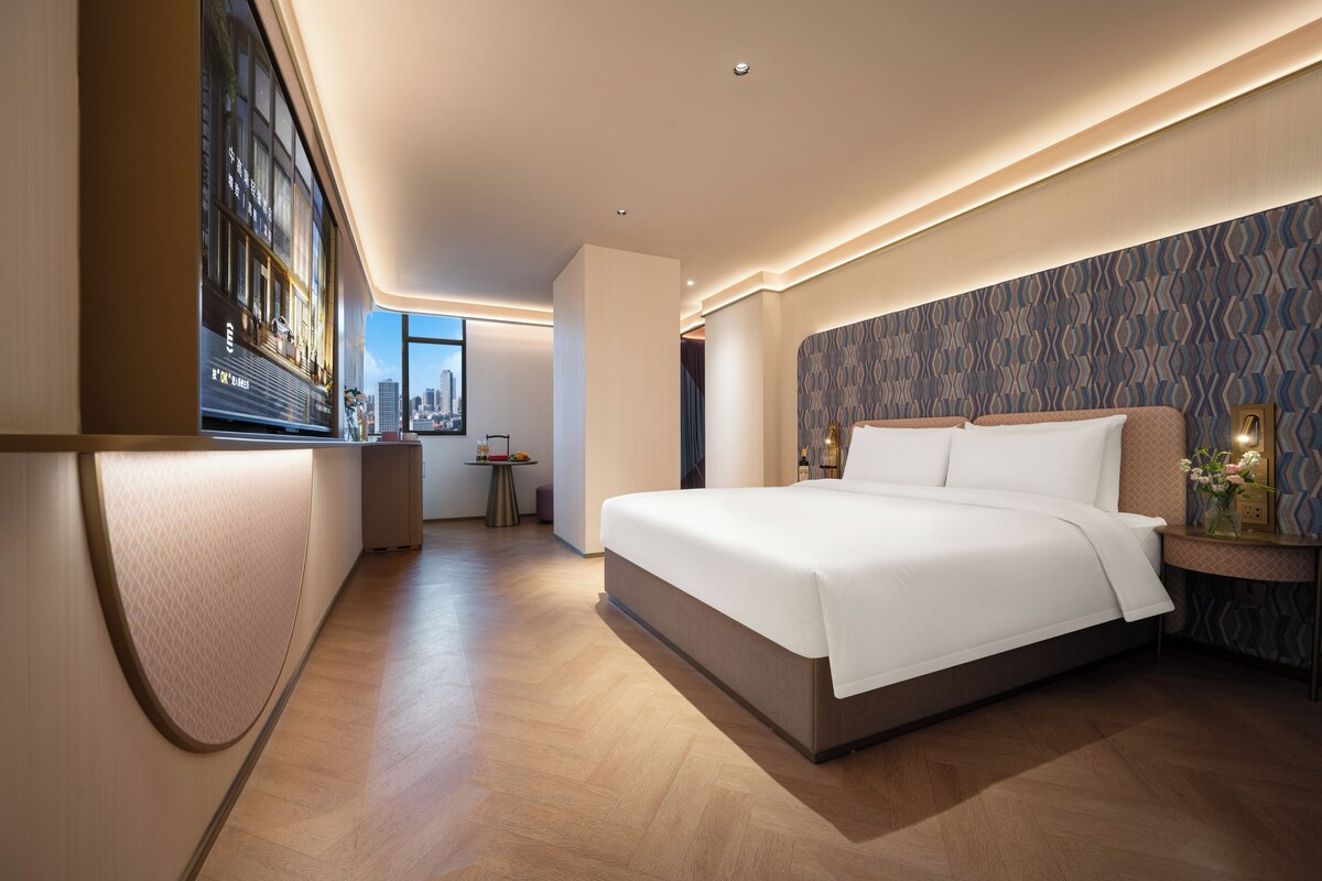 A modern hotel room with a large bed, a desk, and a view of the cityscape.