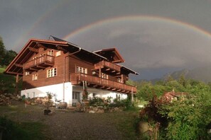 A wooden chalet with a balcony, surrounded by greenery and mountains. - Apartment at Kainzen (Oberammergau)