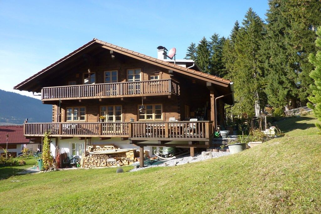 A two-story wooden chalet with a balcony, surrounded by a mountainous landscape.