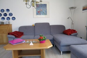 Ein modernes Wohnzimmer mit einem grauen Ecksofa, einem Couchtisch aus Holz, einer rosa Ordner, einer roten Decke und einem Blumentopf.