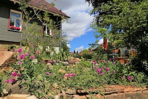 A house with a red-tiled roof, a window with a red frame, and a garden with pink flowers and green plants. - ||--------------------------Old lodging house-----------------------|| (Wernigerode)