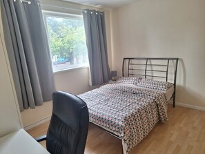 Un lit simple avec un cadre en métal et une literie à motifs, une chaise, une table de chevet et une fenêtre avec des rideaux.