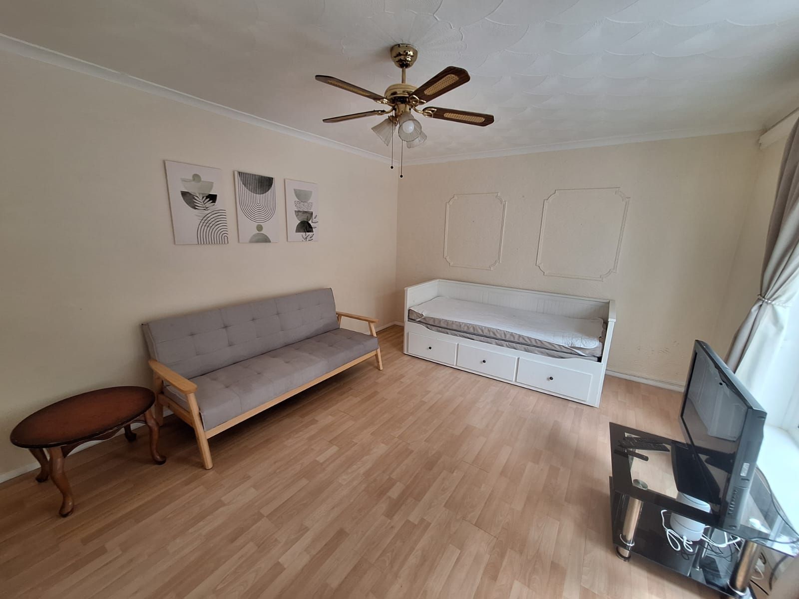 A room with a grey sofa, a bed with storage drawers, a ceiling fan, and a television on a stand.